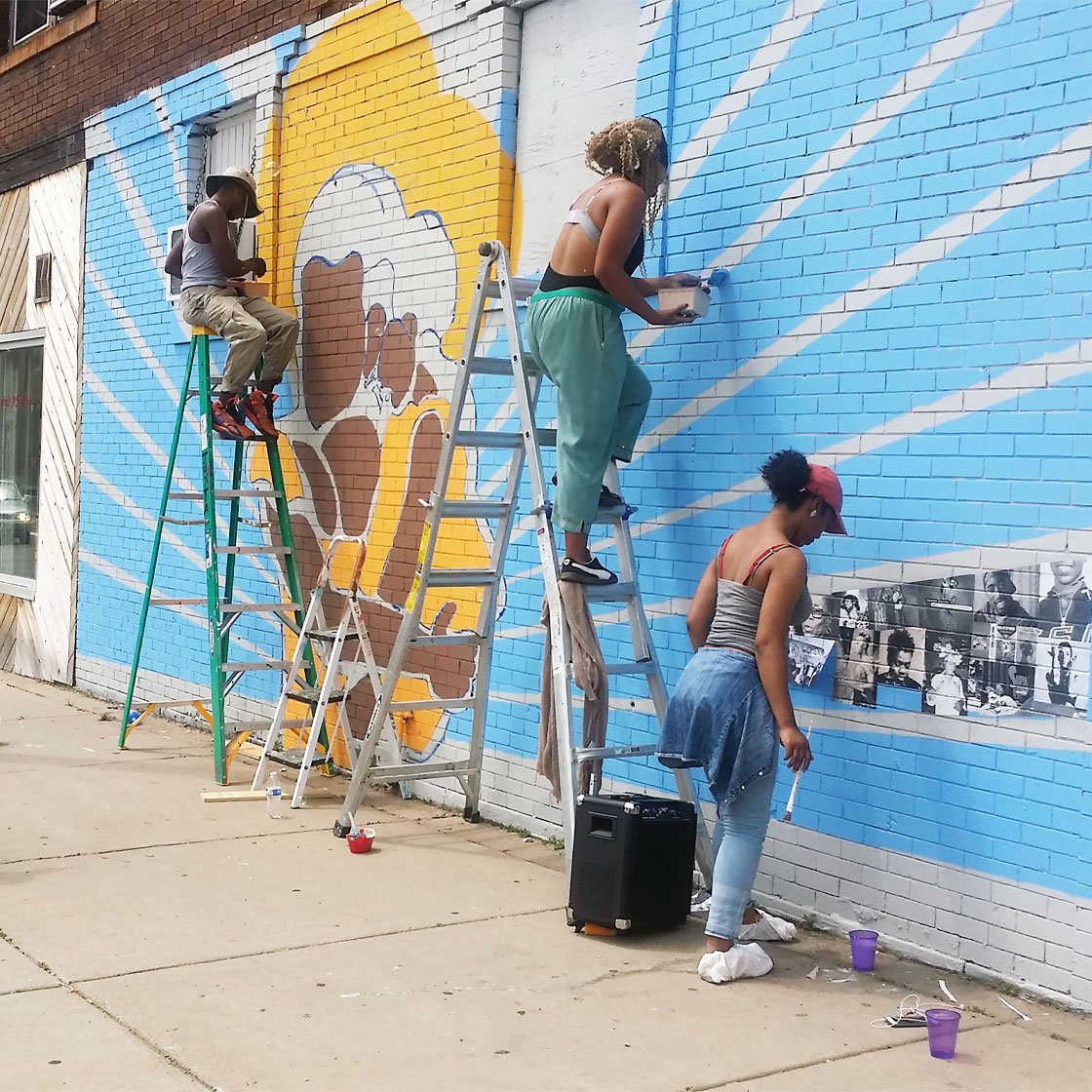 Young people working on an outdoor large-scale mural of a woman holding a child with rays radiating from them