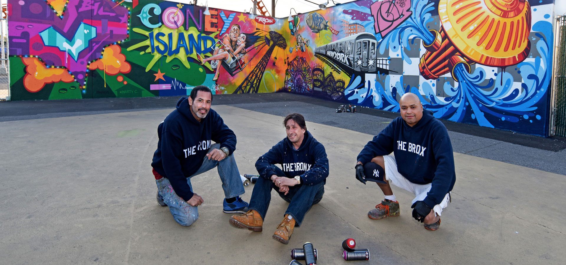 Three people sitting with spray paint cans in front of a colorful mural