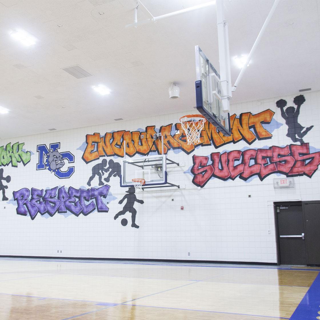 A gymnasium basketball court with a painted wall mural with the words Attitude, Hard Work, Respect, Encouragement, Success