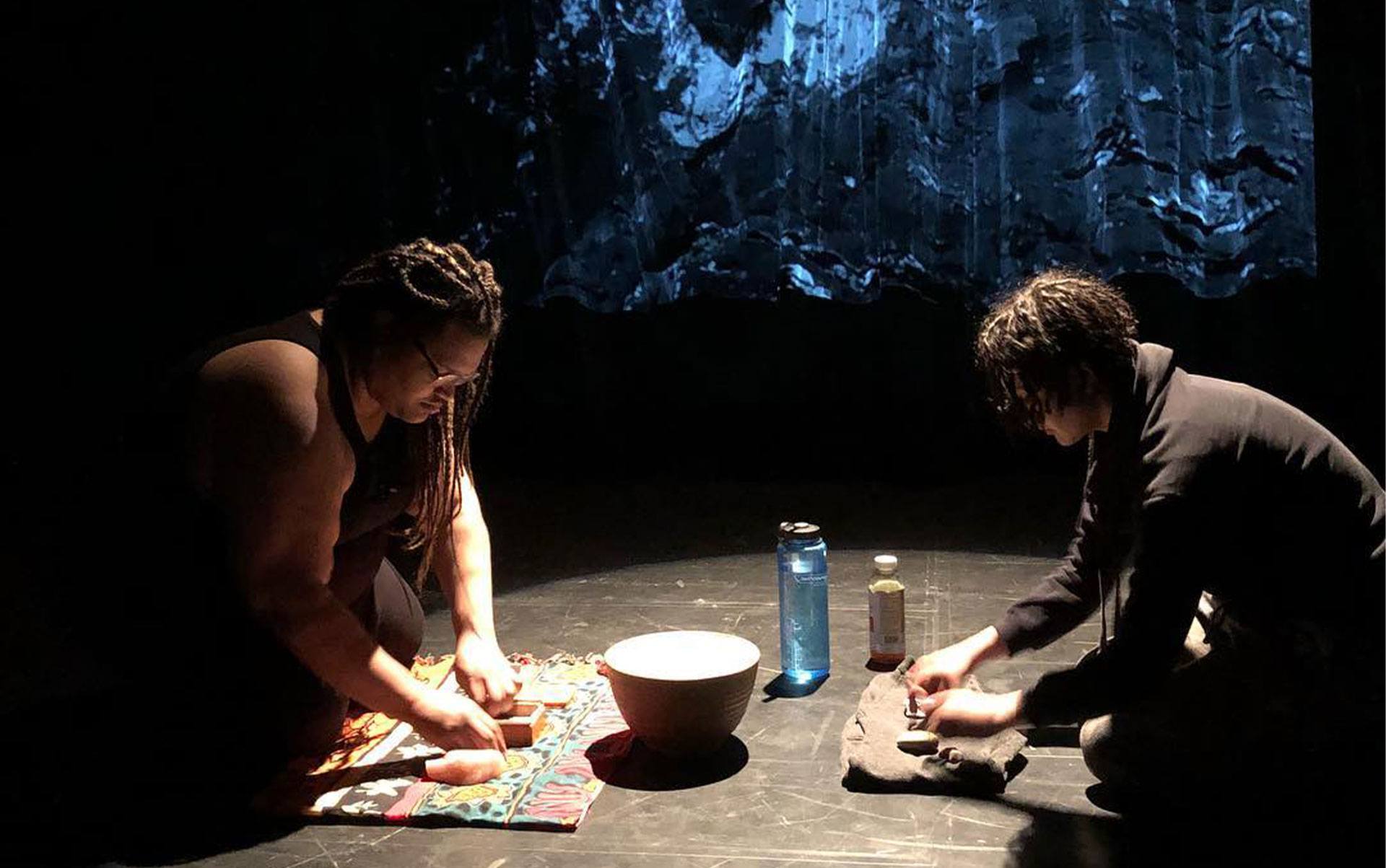2 people sit working on different objects with their hands in a performance on a stage