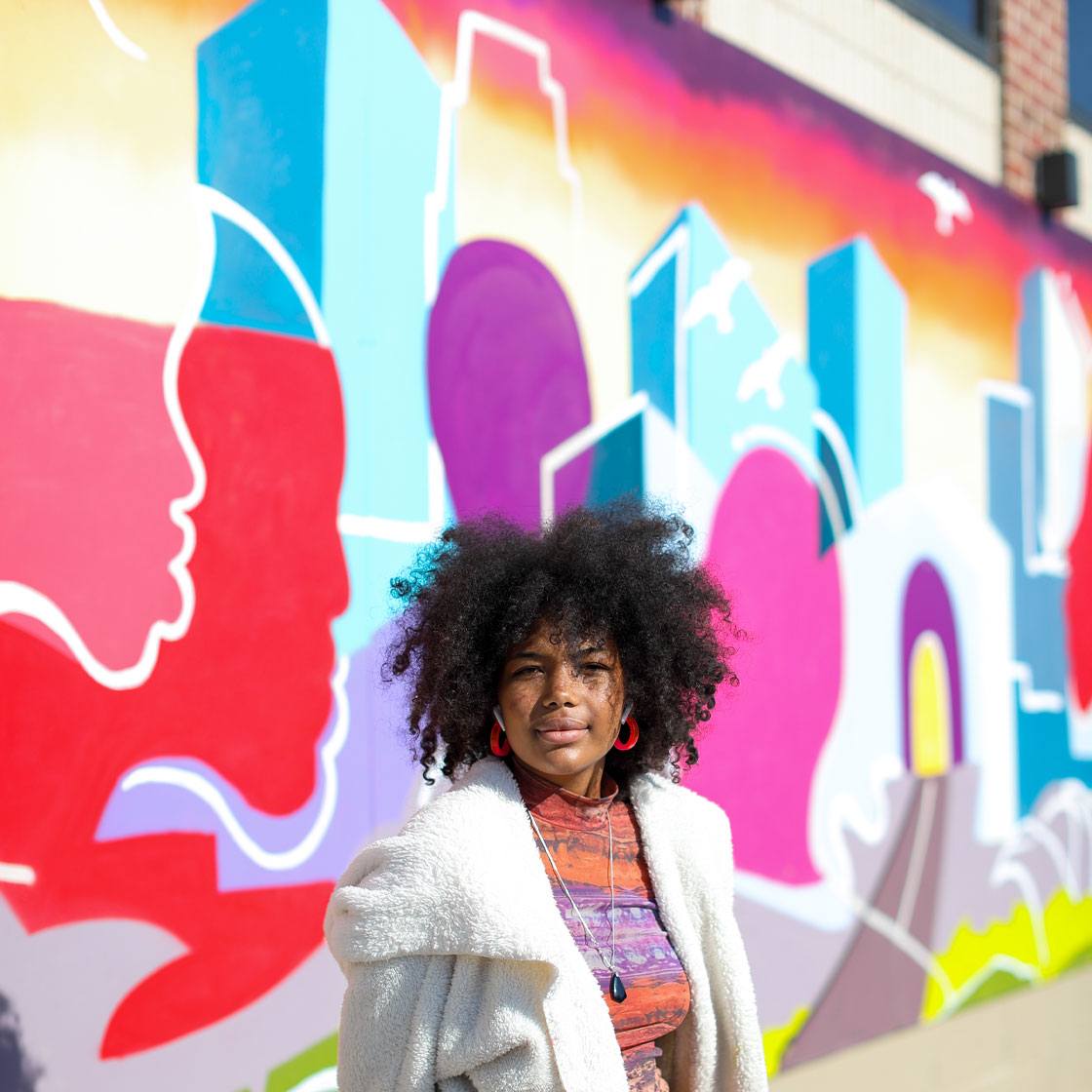 A mural painted with acrylic and spray paints depicting faces and buildings intermingling to represent the city of Minneapolis working together in unity. Apprentice Bereket Weddall poses in front of the mural.